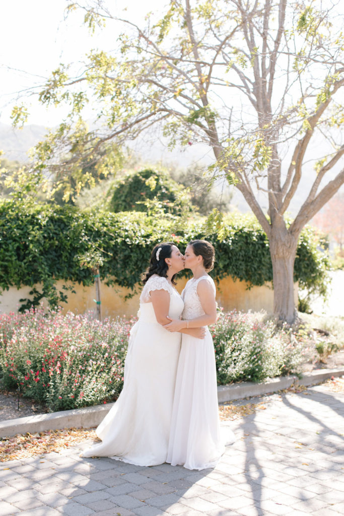 two brides kissing each other in a scenic setting with flowers and trees in carmel