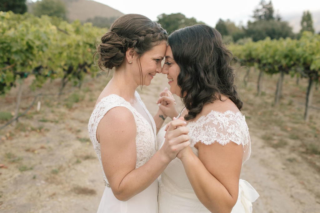 two brides with their heads together smiling and holding hands in a vineyard in Carmel california