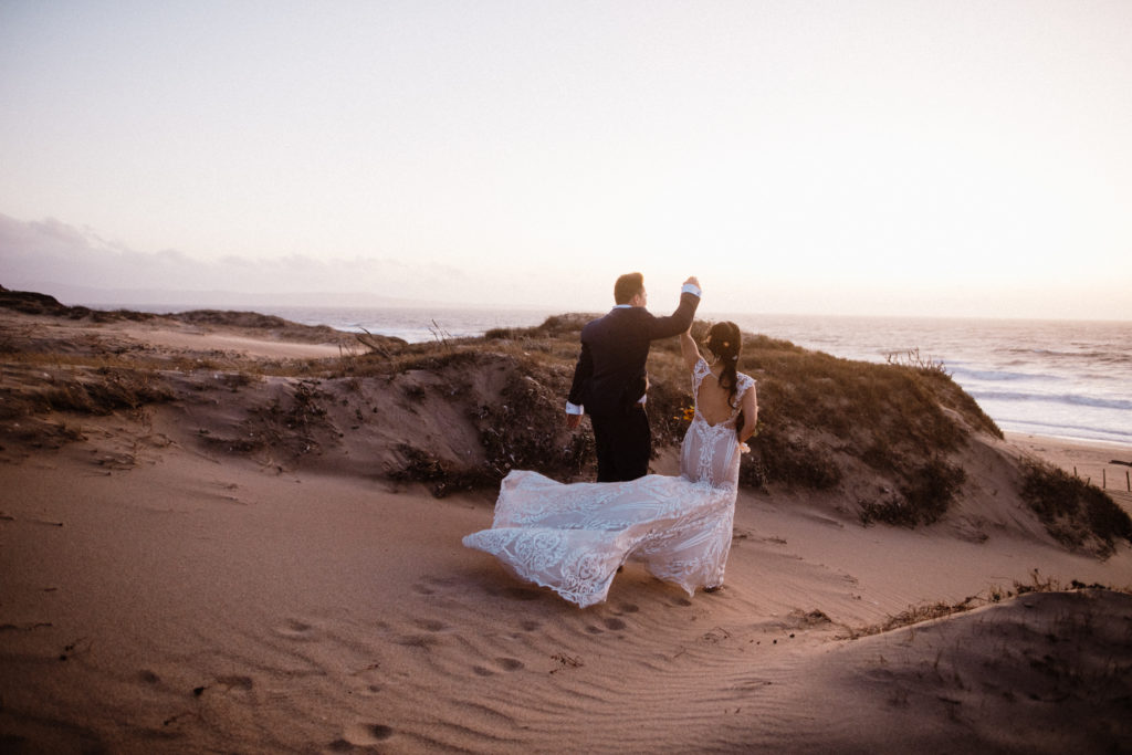 Bride and groom celebrating with arms in the air walking down a rugged beach in california at sunset 